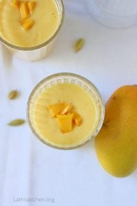 Two glasses of Mango Lassi with a mango on the side and cardamom.
