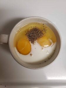 Eggs crack open in a mug with milk and salt and pepper.