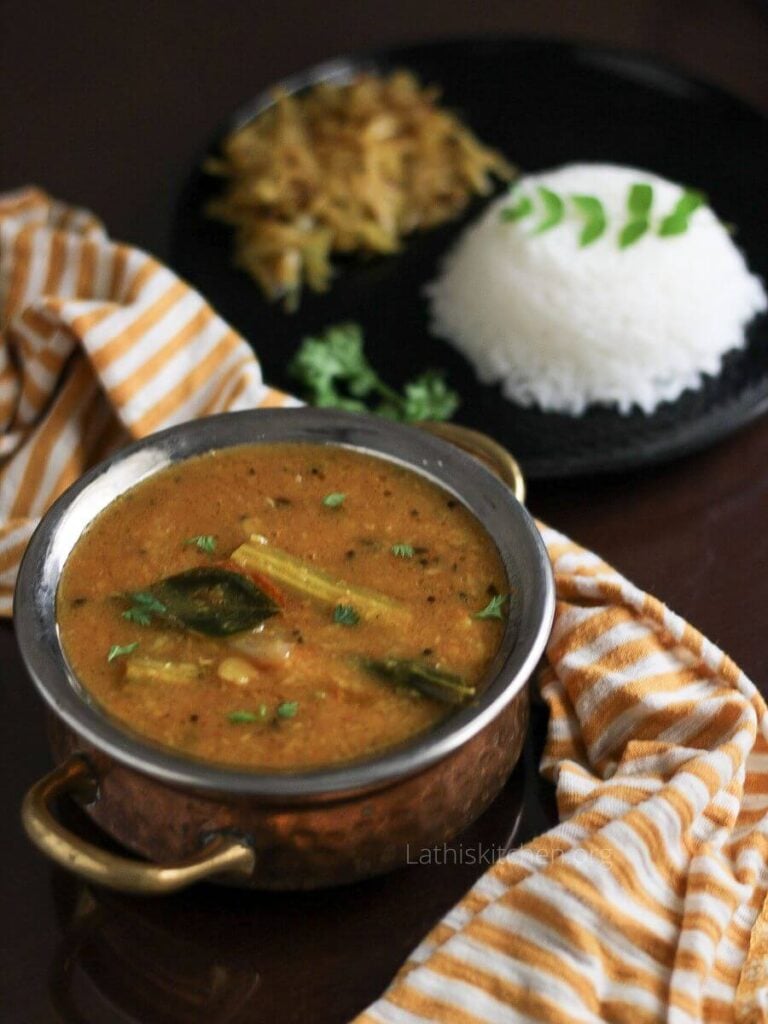 A bowl of sambar in front of a plate with rice and vegetables.