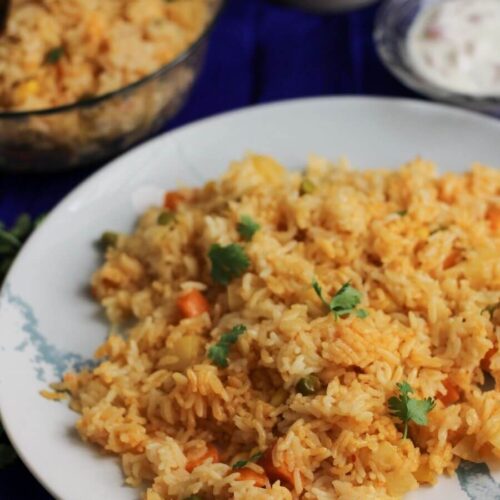 Close up of vegetable biryani in a plate with bowl of rice in out of focus.