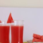 Two glasses filled with watermelon lemonade with pieces of watermelon in a plate and cut lemon wedges.