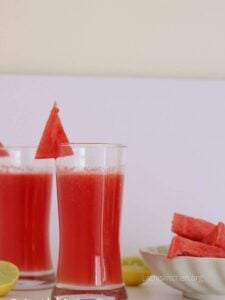 Two glasses filled with watermelon lemonade with pieces of watermelon in a plate and cut lemon wedges.