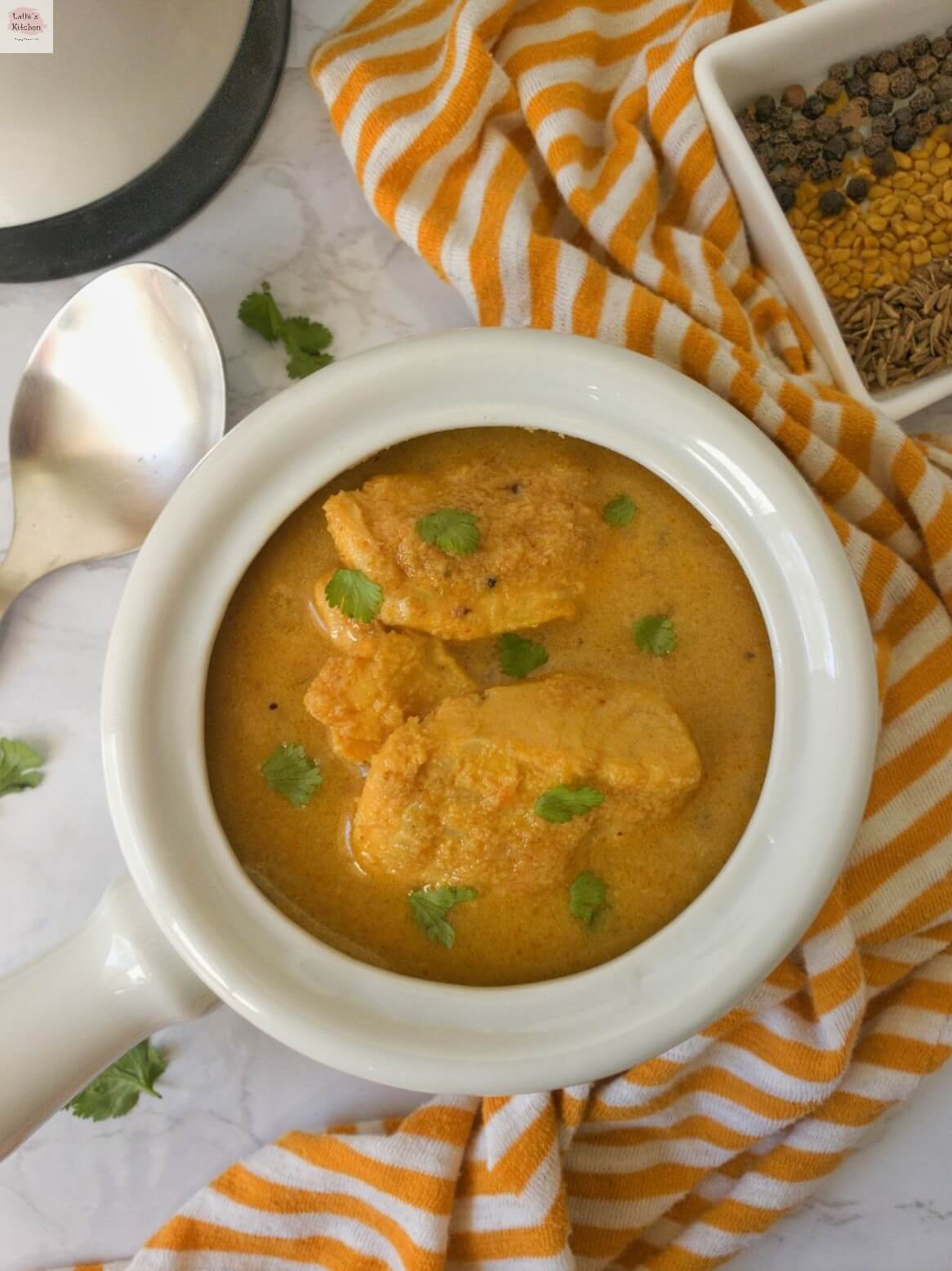 Bowl of kerala fish curry with coconut along with spoon and spices on its side.